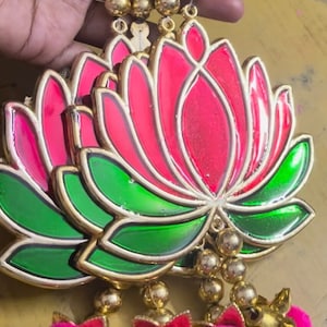 May include: A decorative hanging ornament featuring a layered lotus flower design with pink and green accents. The ornament is adorned with gold beads and pink pom-poms.