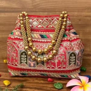Può includere: Una borsa ricamata rossa con manici di perline dorate. La borsa presenta motivi intricati in oro, verde e altri colori. La borsa è decorata con paillettes e ha una forma strutturata, suggerendo che si tratti di una pochette o di una piccola borsa.