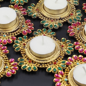 Könnte beinhalten: Eine Sammlung goldfarbener Kerzenhalter mit rosa und grünen Akzenten. Jeder Halter hat eine runde, weiße Kerze und ist mit kleinen, bunten Schmucksteinen verziert.