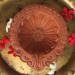 Puede incluir: Una diya de terracota, o lámpara de aceite, con intrincados patrones florales y geométricos tallados. La lámpara está rodeada de flores rojas y blancas, sobre una superficie de latón dorado. La diya es un elemento tradicional utilizado en festivales indios.