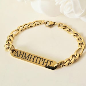 May include: Gold-coloured bracelet featuring a Figaro chain design. A rectangular plate with the name "ΔΗΜΗΤΡΗΣ" in black lettering is centered on the bracelet. The bracelet is displayed on a white surface with a soft, blurred background.