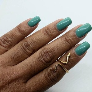 May include: A gold snake ring with a coiled design. The ring is worn on a finger with teal nail polish.