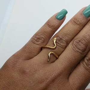 May include: A gold snake ring with a textured finish. The ring is worn on the middle finger of a hand with green nail polish.