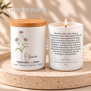 May include: Two white soy candles in glass jars. One has a wooden lid and features a purple aster flower illustration, the name Olivia, and the text September - Aster Love Wisdom Patience Faith. The other is lit and displays a long quote.