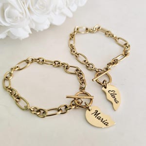 Può includere: Due braccialetti dell'amicizia color oro con maglie a catena ovali. Ogni braccialetto ha una chiusura a levetta e un ciondolo a forma di cuore. Un ciondolo dice "Maria" e l'altro dice "Elena".