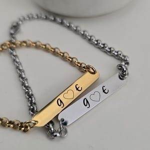 Könnte beinhalten: Zwei passende Armbänder mit einer goldenen und silbernen Kette. Jedes Armband hat ein kleines rechteckiges Schild mit dem Wort "g" und einem Herzsymbol, gefolgt vom Buchstaben "e".