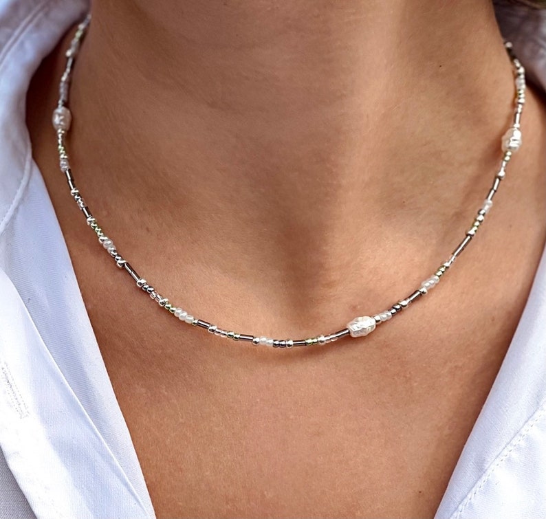 Puede incluir: Un collar de cuentas plateadas y blancas con detalles de perlas. El collar es delicado y presenta peque&ntilde;as cuentas redondas.