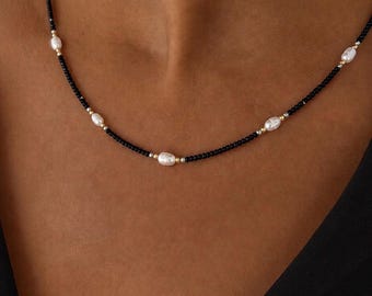 Puede incluir: Un collar de cuentas que presenta pequeñas cuentas de semillas negras intercaladas con perlas de agua dulce ovaladas blancas y diminutas cuentas espaciadoras metálicas de tono dorado. El collar tiene un patrón simple de cuentas negras y perlas.