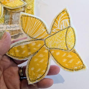 May include: A handmade fabric daffodil with yellow petals and a yellow trumpet, stitched with black thread. The petals have a white and yellow patterned design. The flower is held in a hand, with a glimpse of another fabric art piece in the background.