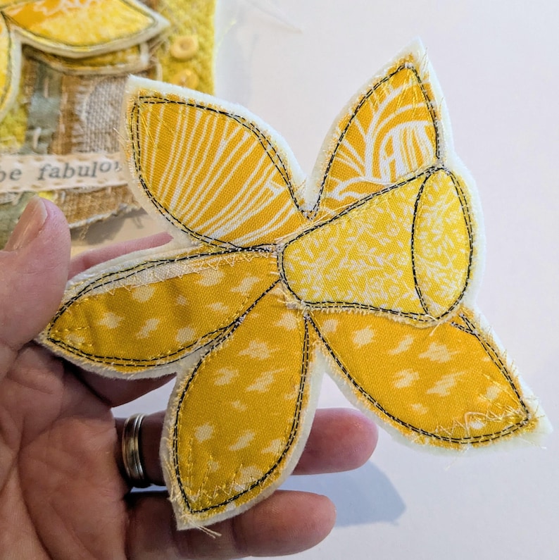 May include: A handmade fabric daffodil with yellow petals and a yellow trumpet, stitched with black thread. The petals have a white and yellow patterned design. The flower is held in a hand, with a glimpse of another fabric art piece in the background.