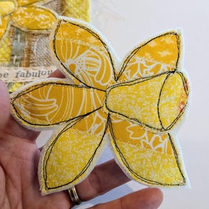May include: A handmade yellow and white fabric flower with black stitching. The flower has six petals and a central trumpet shape. The fabric has a floral pattern. The flower is likely a decorative item or part of a larger craft project.