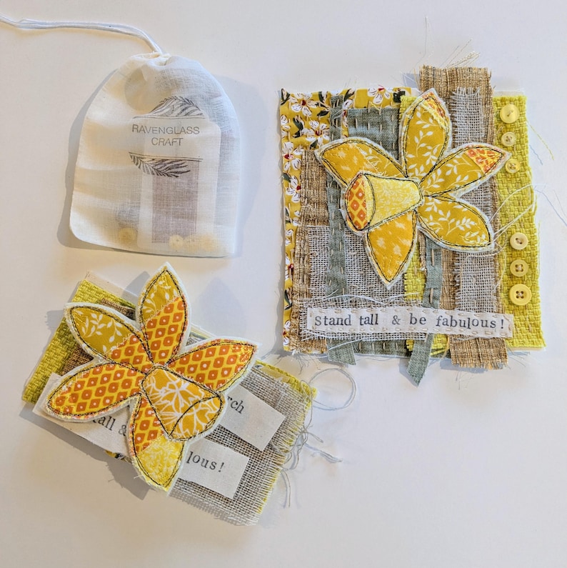 May include: A handmade craft set featuring yellow daffodil fabric art. The set includes a drawstring bag with text, two fabric art pieces with the text "Stand tall & be fabulous!" and "March", and a daffodil design.