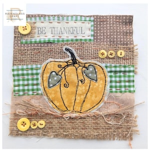 May include: A fabric wall hanging featuring a central yellow pumpkin appliqué with black stitched details and two heart-shaped leaves. The background consists of layered hessian and green gingham fabric strips with the text BE THANKFUL and yellow buttons.