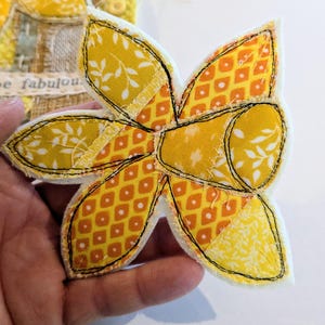 May include: A handmade fabric daffodil flower with yellow and orange patterned petals and a yellow center. The flower is outlined with black stitching and held in a hand. The flower is a craft item.