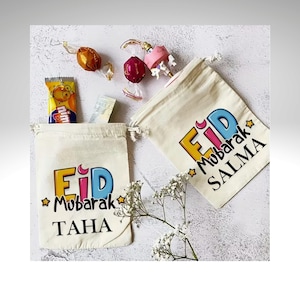 May include: Two off-white drawstring bags with the words "Eid Mubarak" in yellow and blue, and names "TAHA" and "SALMA" in black. The bags are filled with candy and sweets. Scattered around are wrapped candies and a small yellow candy wrapper.