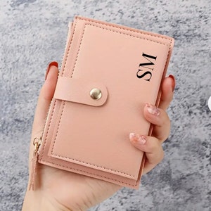 May include: A blush pink bi-fold wallet with a snap closure and a gold zipper. The wallet has the initials "SM" printed on the front. The wallet is held in a hand.