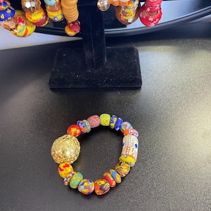 Puede incluir: Una colorida pulsera de cuentas con una gran cuenta esférica dorada y varias cuentas estampadas en rojo, amarillo, azul y verde. La pulsera se exhibe sobre una superficie negra, mostrando su vibrante diseño.