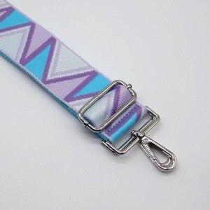 Könnte beinhalten: Ein farbenfroher Taschenriemen mit Zickzackmuster in Blau-, Lila- und Weißtönen. Der Riemen hat silberfarbene Metallbeschläge, darunter ein Karabinerhaken und verstellbare Schnallen. Für Handtaschen.