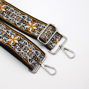 May include: Two black and gold embroidered bag straps with white floral and geometric patterns. The straps have silver metal swivel hooks and rectangular metal adjusters. The design features a repeating pattern of stylized flowers and swirls.
