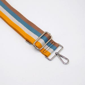 May include: A striped bag strap in yellow, white, blue, and brown. The strap features silver-coloured metal hardware, including a rectangular buckle and a swivel clasp. The strap is angled across a white background.