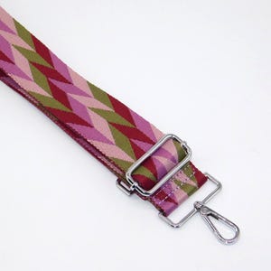 May include: A patterned bag strap featuring a geometric design in pink, green, and burgundy. It has silver-tone metal hardware, including a rectangular buckle and a swivel clasp.