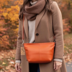 May include: An orange leather crossbody bag with a long strap. The bag is rectangular with a smooth surface and is being worn over a tan coat. The background features fall foliage with red, orange, and yellow leaves.