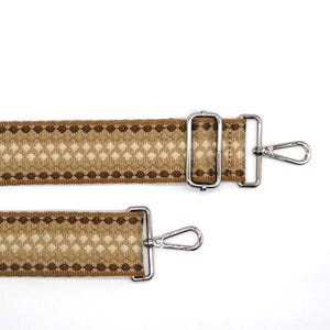 May include: Two brown and beige patterned straps with silver-coloured metal clasps. The straps feature a repeating diamond and dot design. The clasps are rectangular and have a swivel hook for attachment.