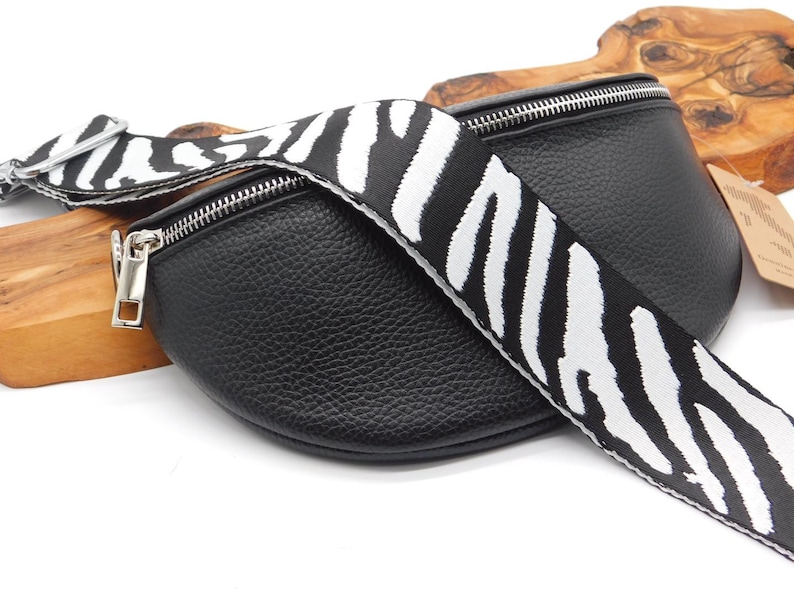 May include: Black leather bum bag with a zebra print strap. The bag features a silver zip and hardware. The strap has a striking black and white zebra stripe pattern. The bag is curved in shape.