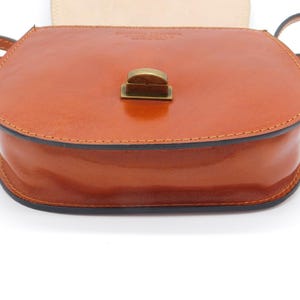 May include: A tan leather crossbody bag with a rounded shape and a flap closure. The bag has a shiny finish, a black trim, and a brass-coloured clasp. The strap is thin and tan. The bag is likely for carrying personal items.