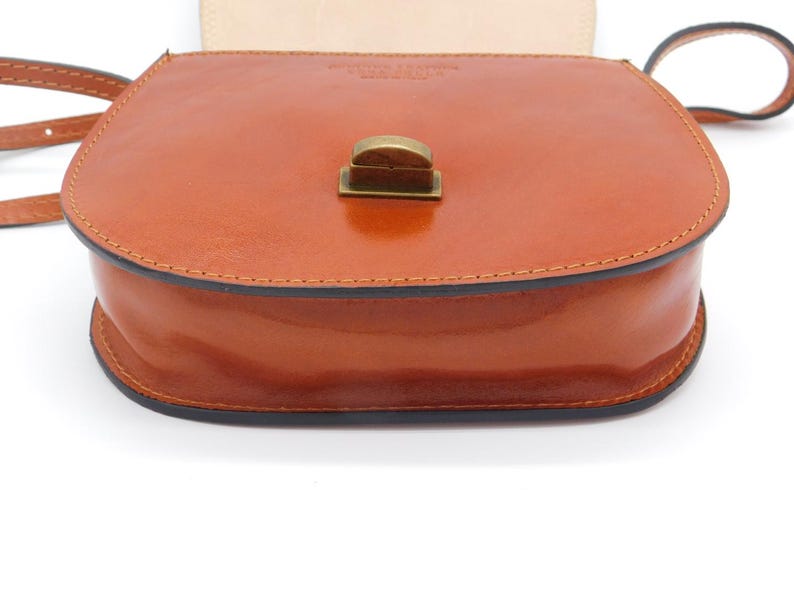 May include: A tan leather crossbody bag with a rounded shape and a flap closure. The bag has a shiny finish, a black trim, and a brass-coloured clasp. The strap is thin and tan. The bag is likely for carrying personal items.