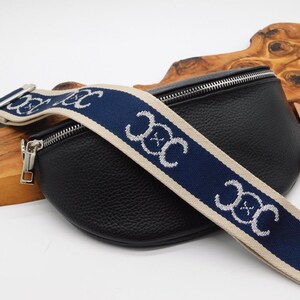 May include: Black leather bum bag with a navy blue and beige patterned strap. The strap features a repeating white design. The bag has a silver zip and hardware. The bag is on a wooden surface.