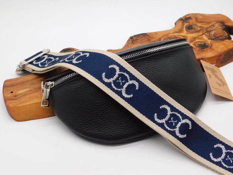 May include: Black leather bum bag with a navy blue and beige patterned strap. The strap features a repeating white design. The bag has a silver zip and hardware. The bag is on a wooden surface.
