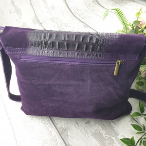 May include: A deep purple suede crossbody bag with a dark, textured flap. The bag features a purple zipper and a gold-colored zipper pull. The strap is also purple. The bag is a stylish accessory.