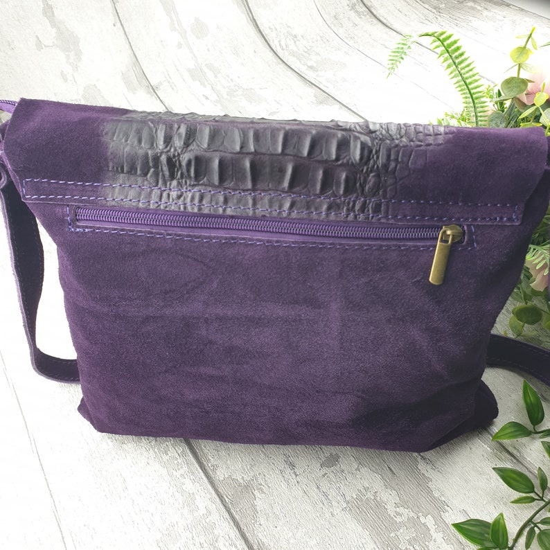 May include: A deep purple suede crossbody bag with a dark, textured flap. The bag features a purple zipper and a gold-colored zipper pull. The strap is also purple. The bag is a stylish accessory.
