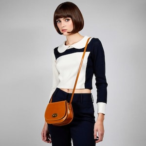 May include: A tan leather crossbody bag with a curved design and a gold clasp. The bag has a long, thin strap. The person is wearing a navy and white long-sleeved top and navy trousers.