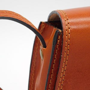 May include: Close-up of a tan leather bag with a curved handle. The bag has visible stitching in a contrasting colour, and a metal rivet. The leather has a smooth, polished appearance, suggesting a high-quality product.