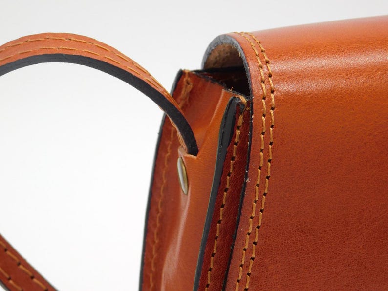May include: Close-up of a tan leather bag with a curved handle. The bag has visible stitching in a contrasting colour, and a metal rivet. The leather has a smooth, polished appearance, suggesting a high-quality product.