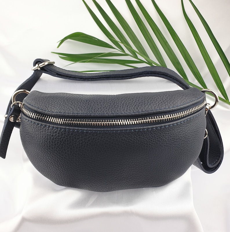 May include: A navy blue leather bum bag with a silver zip and metal hardware. The bag has a curved shape and a strap for wearing around the waist or across the body. The leather has a textured appearance.