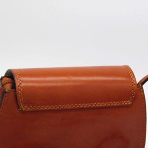 May include: A close-up of a brown leather crossbody bag. The bag has a flap closure with visible stitching and a shoulder strap. The leather has a glossy finish.