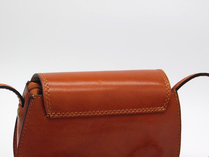 May include: A close-up of a brown leather crossbody bag. The bag has a flap closure with visible stitching and a shoulder strap. The leather has a glossy finish.