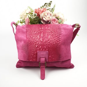 May include: A fuchsia leather shoulder bag with a flap and a crocodile-skin texture. The bag has a suede body and a buckle closure. A bouquet of pink and white flowers sits on top of the bag.