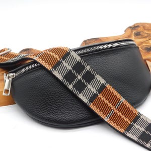 May include: Black leather bum bag with a plaid strap in black, white, and orange. The bag features a silver zip and hardware. The strap is adjustable and can be worn around the waist or across the body.