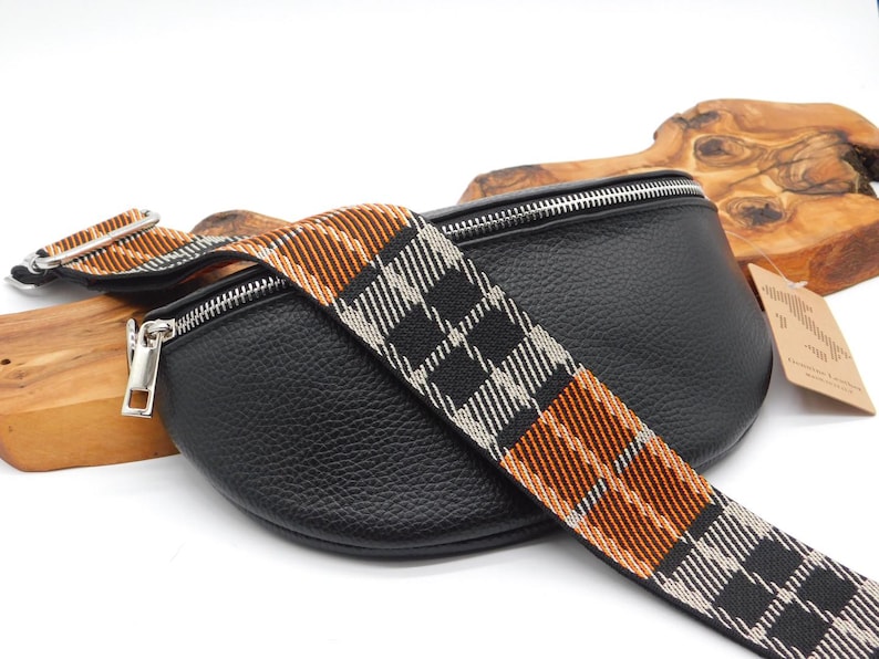 May include: Black leather bum bag with a plaid strap in black, white, and orange. The bag features a silver zip and hardware. The strap is adjustable and can be worn around the waist or across the body.