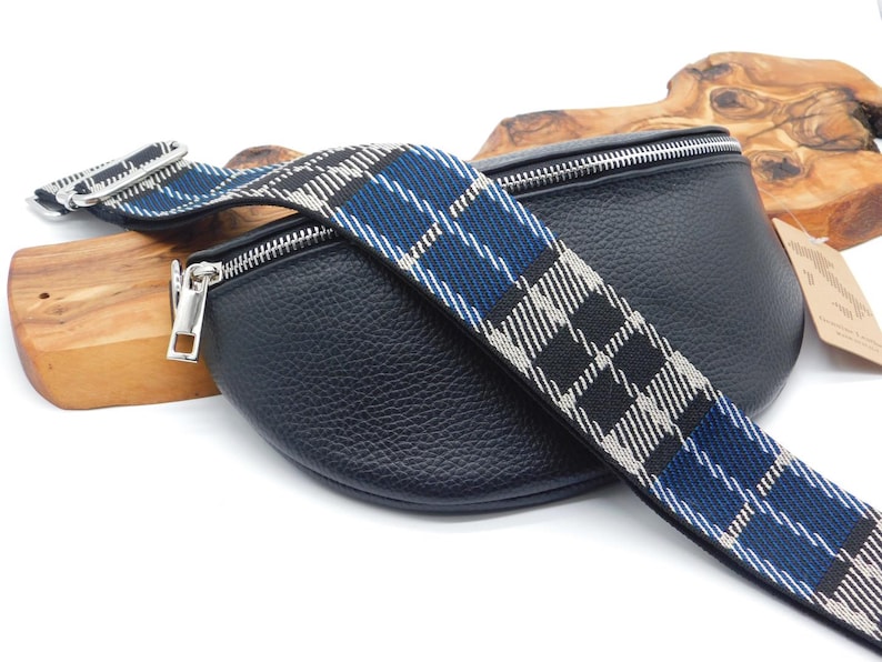 May include: Black leather bum bag with a silver zip and a blue, black, and white tartan strap. The strap is adjustable and has silver hardware. The bag is on a wooden surface.