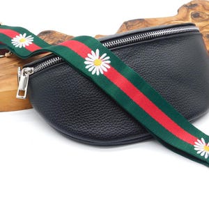 May include: Black leather bum bag with a daisy-patterned strap. The strap has a green background with red stripes and white daisy flowers. The bag features a silver zip and hardware.