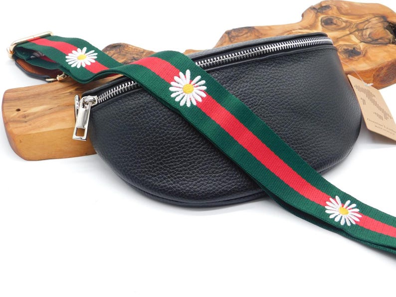 May include: Black leather bum bag with a daisy-patterned strap. The strap has a green background with red stripes and white daisy flowers. The bag features a silver zip and hardware.
