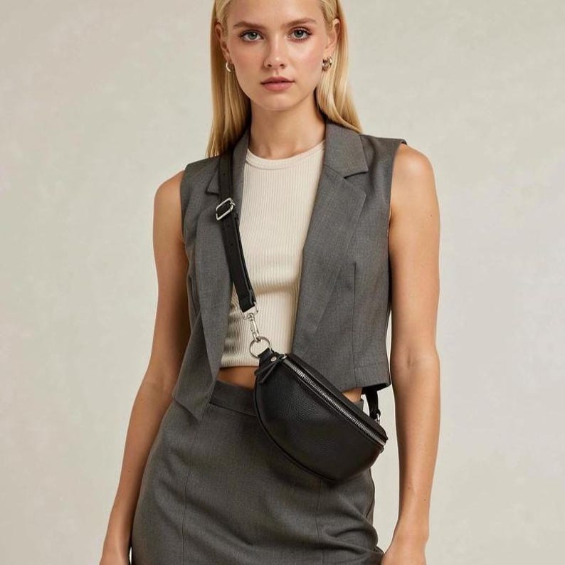 May include: A black leather bum bag with a silver zip is worn across the body. The bag is paired with a grey waistcoat and skirt. The outfit includes a cream top. The bum bag has a black strap with a silver buckle.