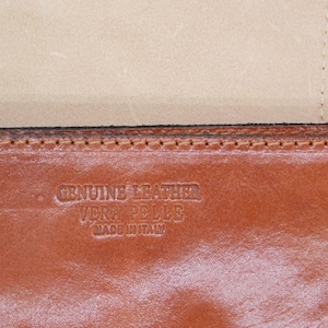 May include: Close-up of a brown leather item with the embossed text "GENUINE LEATHER VERA PELLE MADE IN ITALY". The leather has a rich, warm tone, and the stitching is visible along the edge. The item appears to be a wallet or similar accessory.