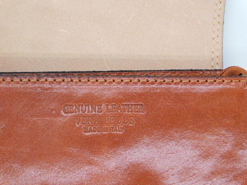 May include: Close-up of a brown leather item with the embossed text "GENUINE LEATHER VERA PELLE MADE IN ITALY". The leather has a rich, warm tone, and the stitching is visible along the edge. The item appears to be a wallet or similar accessory.