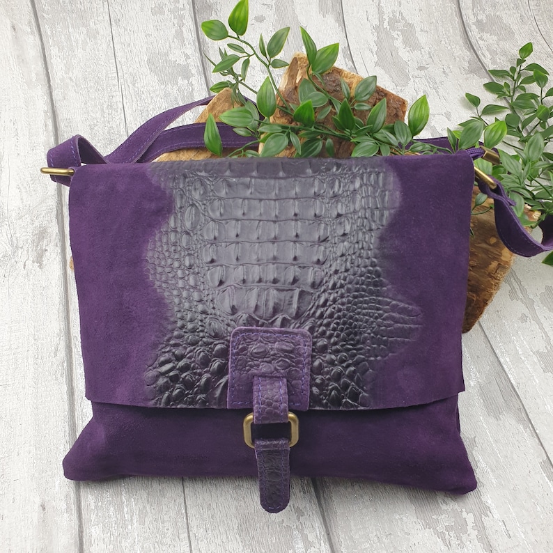 May include: A purple suede crossbody bag with a flap closure. The flap features a dark gray crocodile-textured leather panel. The bag has a matching purple strap and a gold-colored buckle. The bag is designed for everyday use.